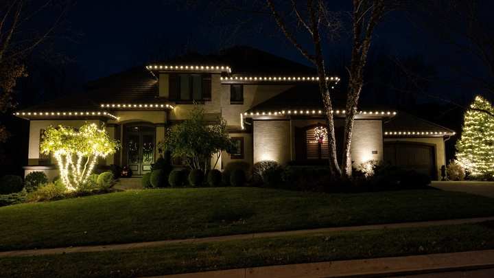 Ranch home at night with warm white roofline lights and lit trees and shrubs